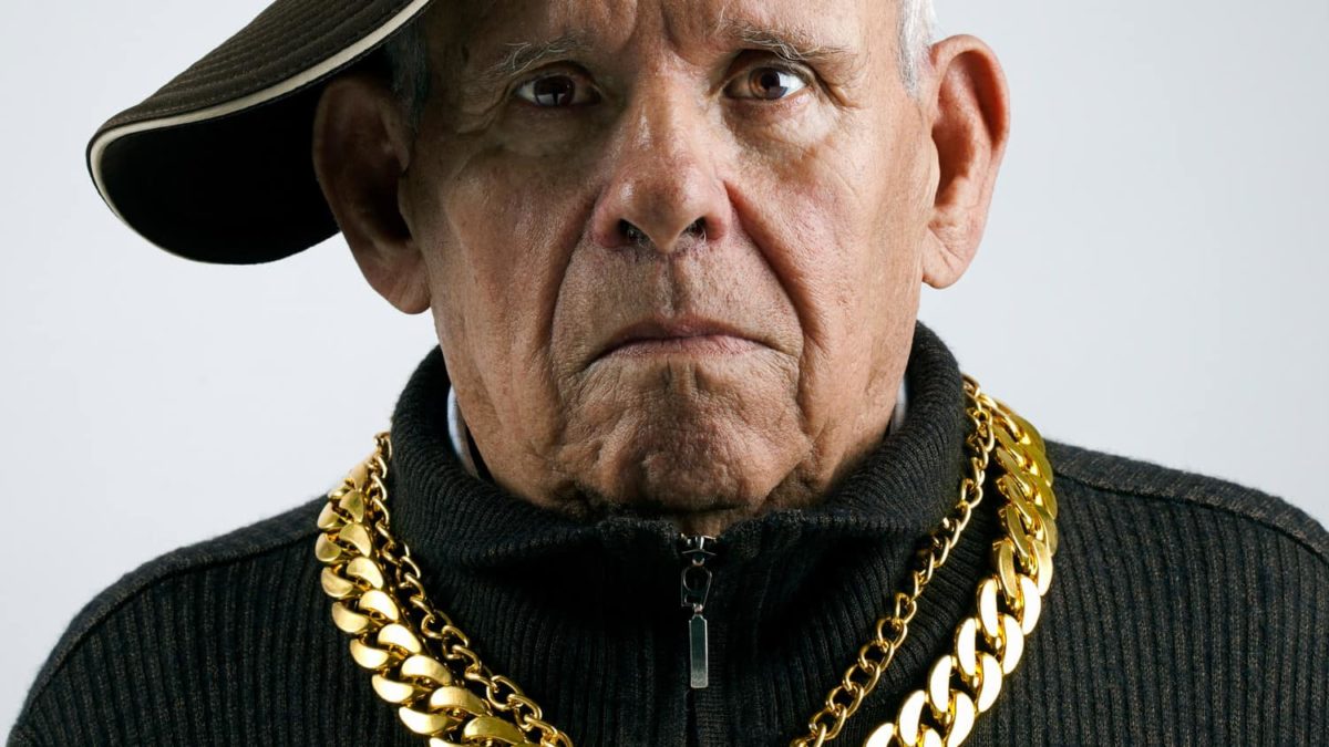 an older man wearing thick gold chains and a baseball cap on the side looks glumly at the camera.