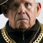 an older man wearing thick gold chains and a baseball cap on the side looks glumly at the camera.