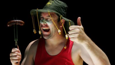 an ocker australian wearing zinc cream on his face and an australian cricket hat with corks and holding a burnt sausage on a fork gives the thumbs up.