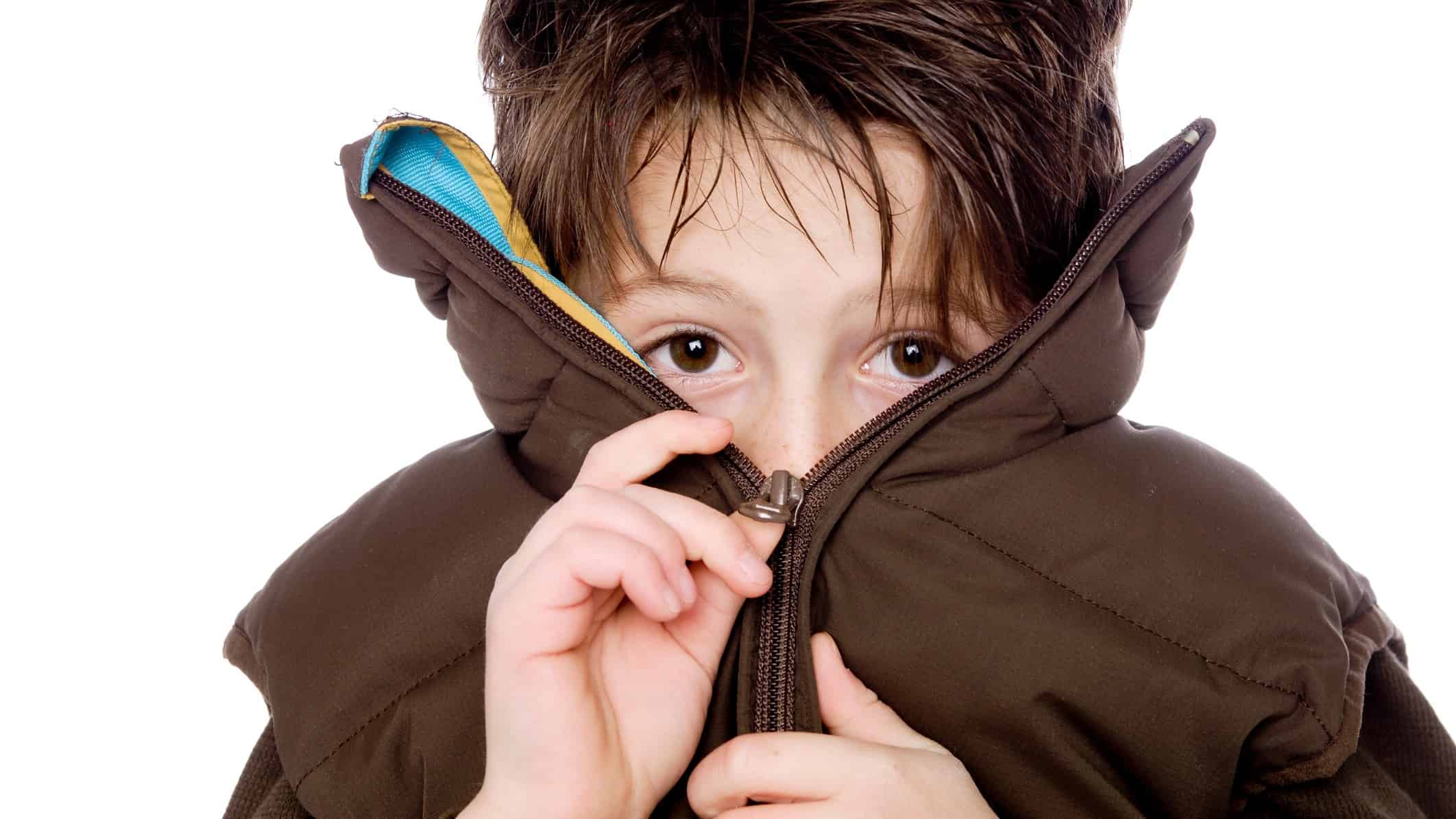 a boy with sad eyes pulls the zip over his mouth and nose while doing up a large jacket where the collar stands up at head height.