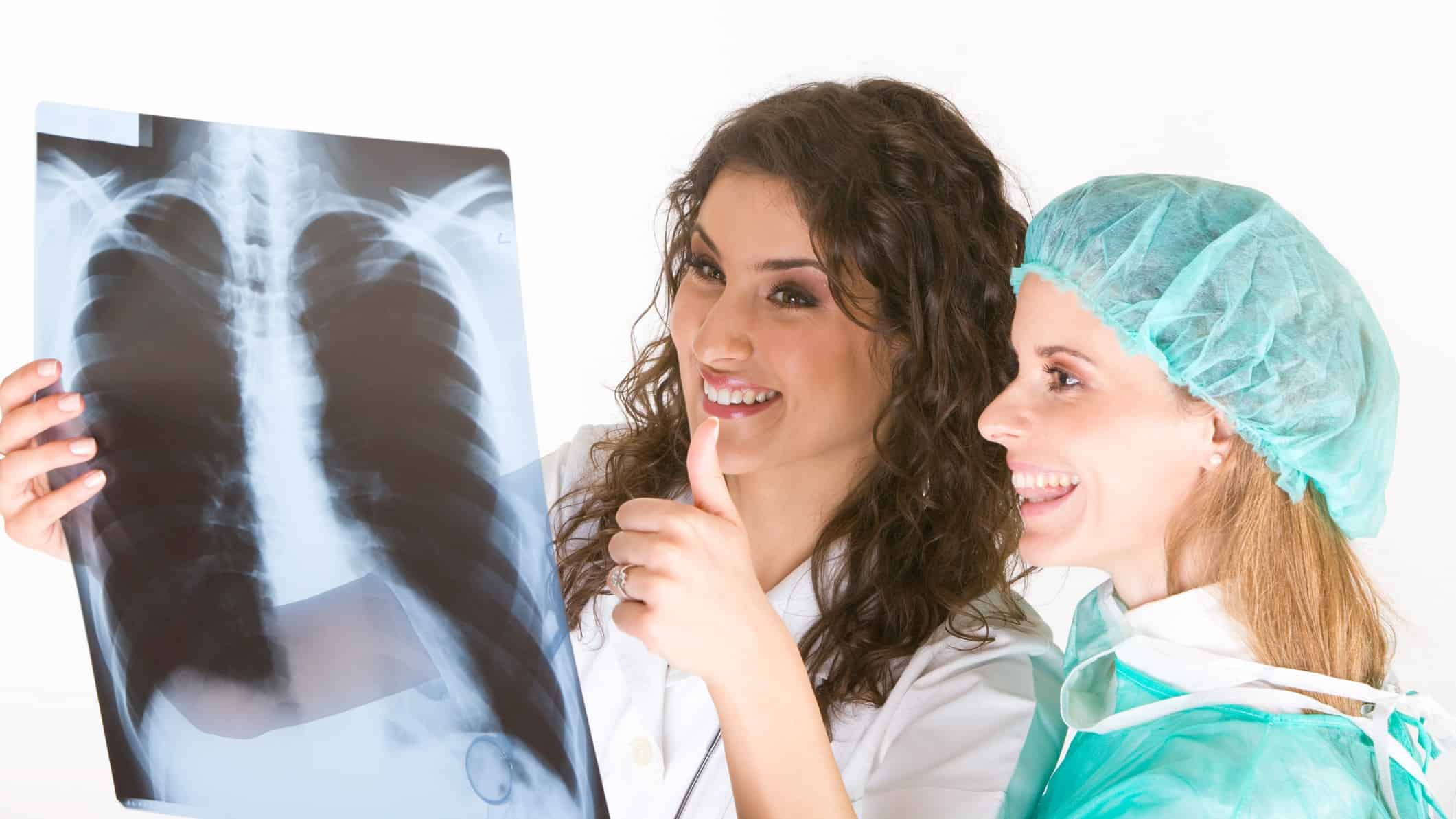 two women, one in a white coat and the other in medical protective gear including a hair cover, mask around her neck and a gown, look happily at an X-ray of a person's chest with one giving the thumbs up sign.
