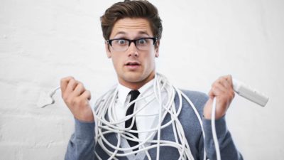 a man wearing a business suit has a wide-eyed surprised expression on his face with a powerboard in one hand and a power plug in the other and yards of power chord wrapped many times over around his neck and body.