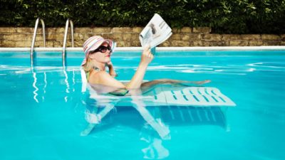 A woman wearing a hat, sunglasses and a bathing suit reads the newspaper while sitting on a lounging chair that's placed in a pool in a relaxing setting.