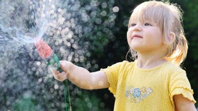 a very young girl looks up at a splash of water coming from a hose that she is holding with the sunlight shining through the water drops.