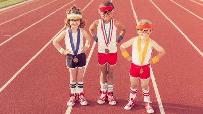 Three children wearing athletic short and singlets stand side by side on a running track wearing medals around their necks and standing with their hands on their hips.