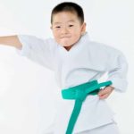 A small child in a judo outfit with a green belt strikes a martial arts pose with his hand thrust forward.