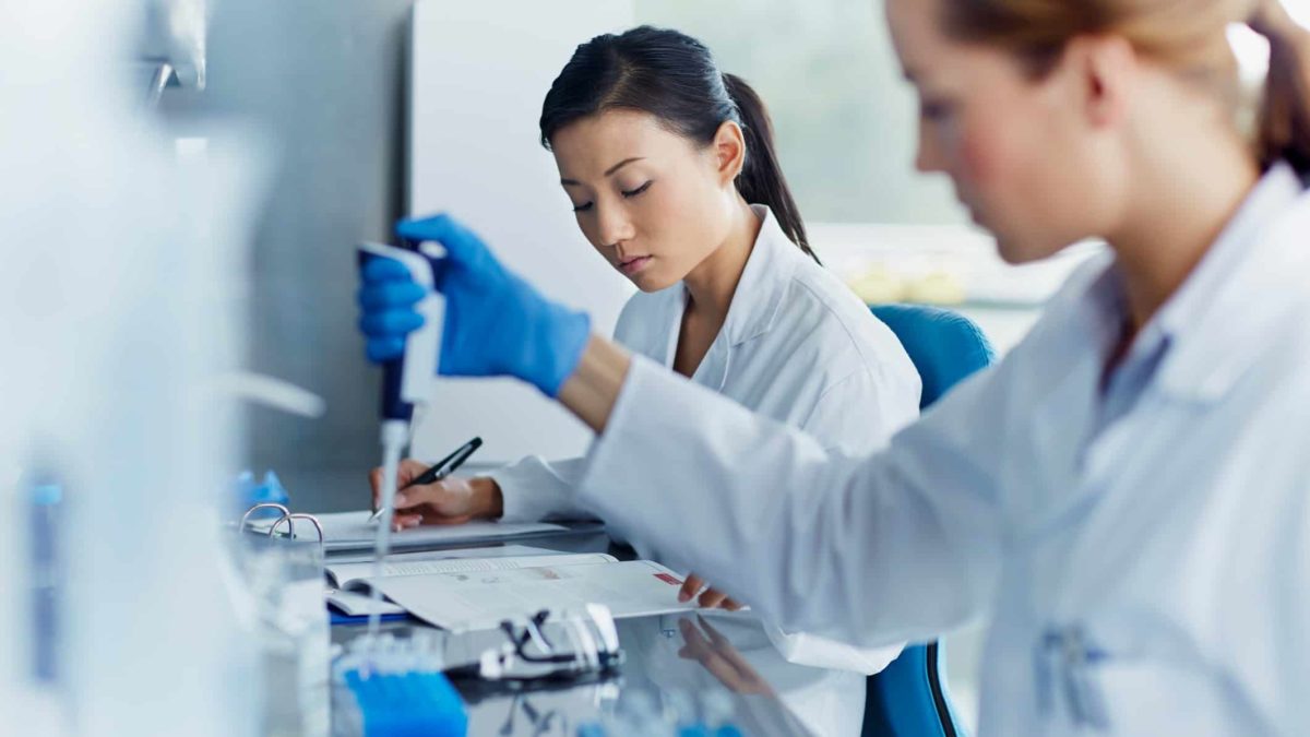 Two laboratory researchers in white coats and gloves sit side by side with scientific equipment and a computer screen conducting medical related research.