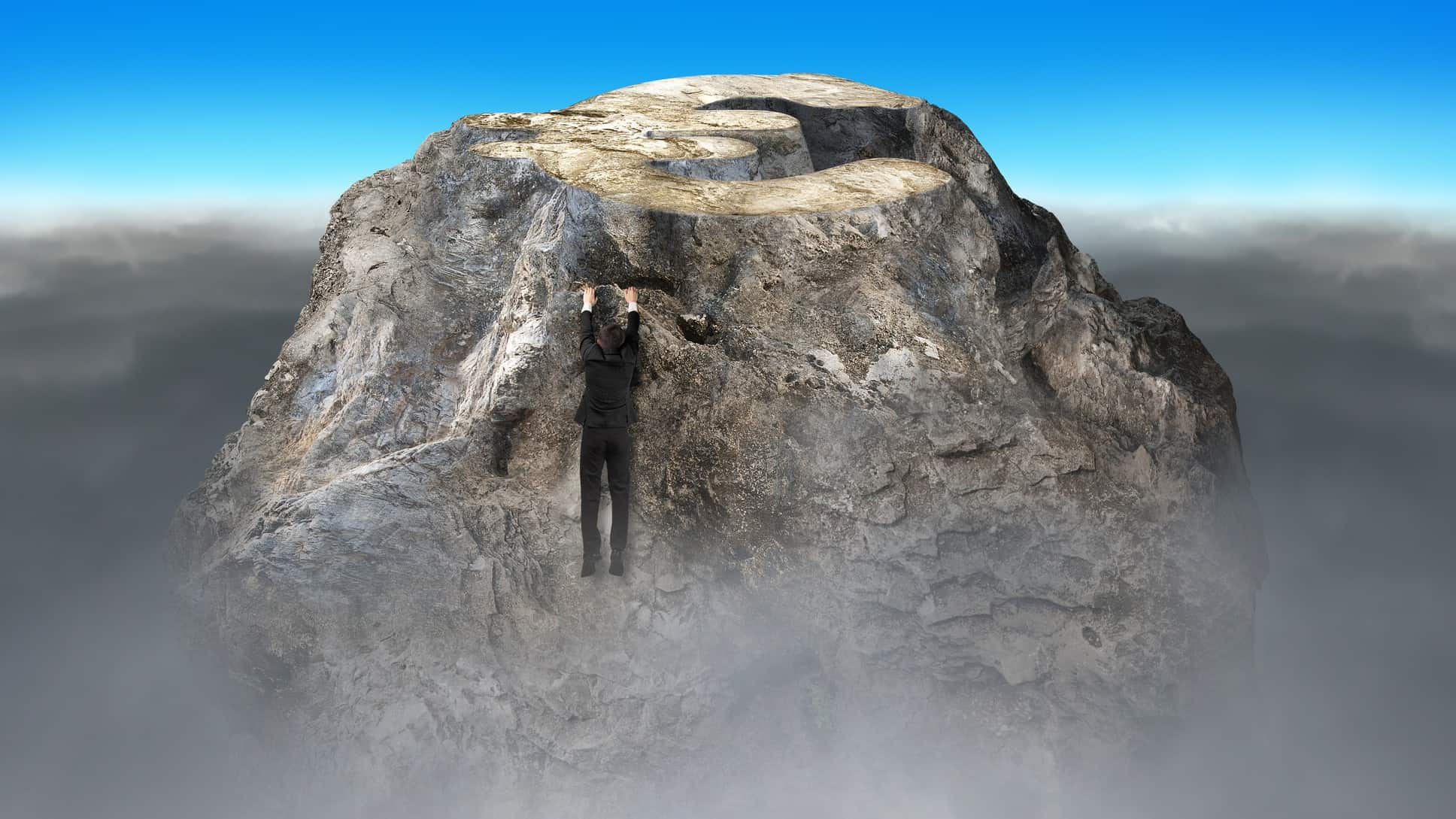a man in a business suit hangs on with his bare hands as he nears the top of a rocky mountain with little footholds and mist swirling around the mountain top.