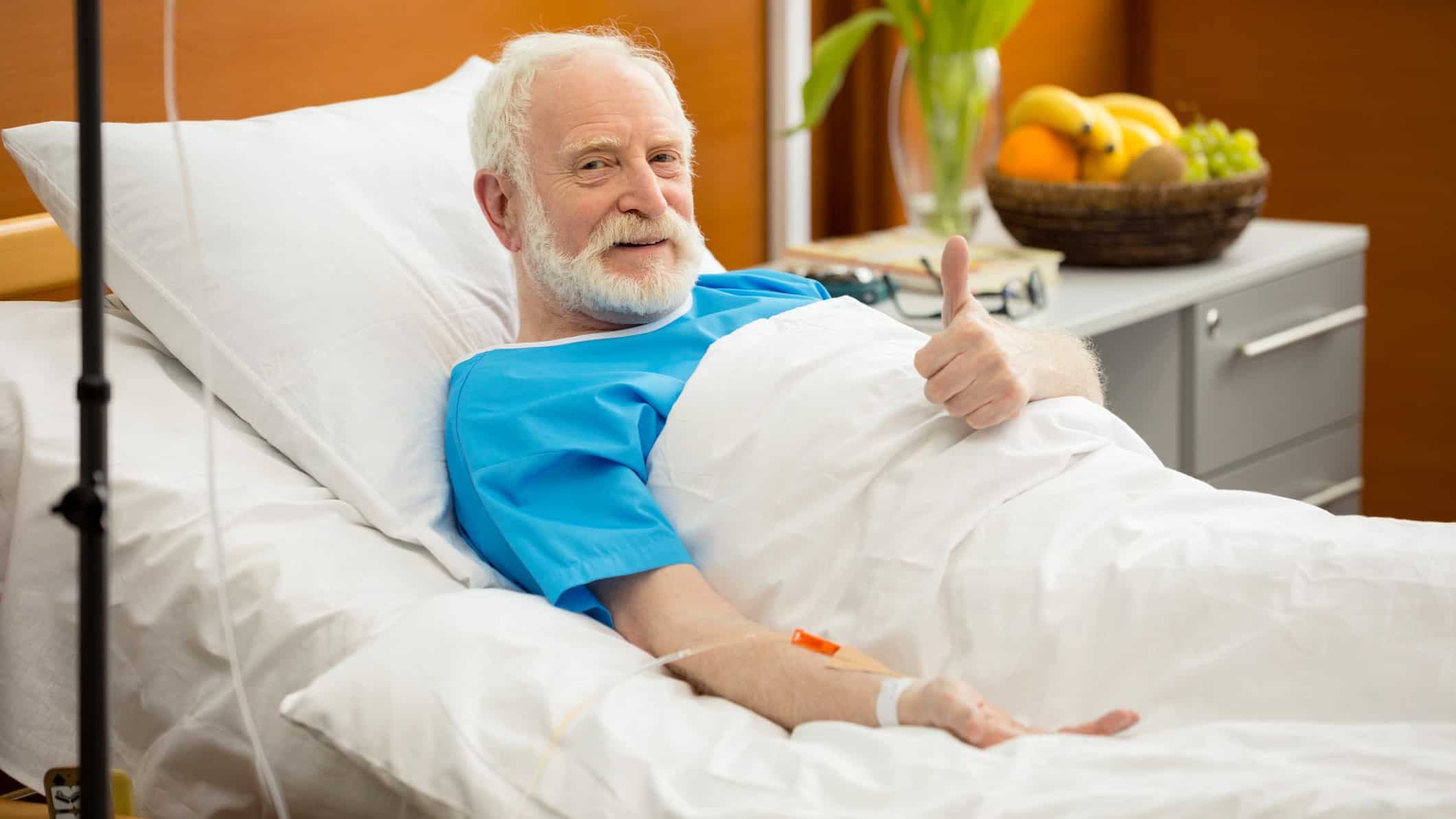 a man in a hospital bed on a drip gives a thumbs up sign while reclining with a bedside table containing a plant and fruit in the background.