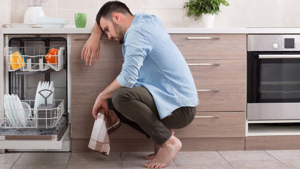 a man slumps to the floor next to his dishwasher with his head resting on his forearm on the kitchen bench and a tea towel in hand. He is looking despondent and sad.