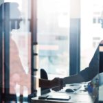 two business people shake hands through the glass wall of a business office with a board table and laptop computer in view between them.