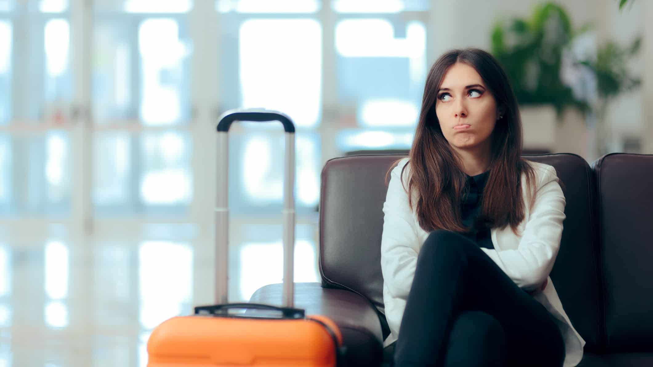 a woman sits next to her wheel along suitcase with the handle raised in a desserted airport with her arms folded and a frustrated, sad expression on her face.
