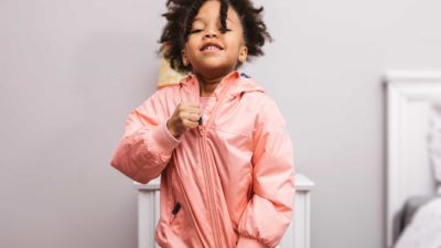 A cute young girl stands with her chest thrust out as she zips up the zip of a shiny pink jacket she is wearing.