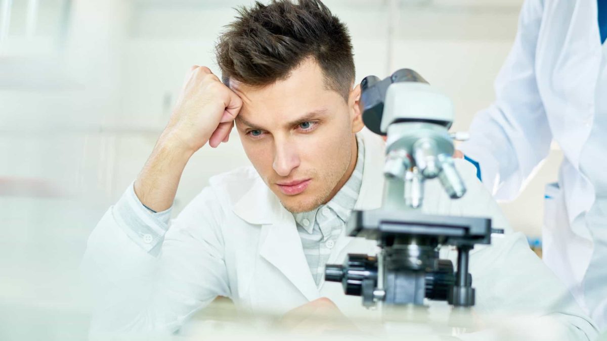A medical researcher rests his forehead on his fist with a dejected look on his face while sitting behind a scientific microscope with another researcher's hand on his shoulder as if giving comfort.