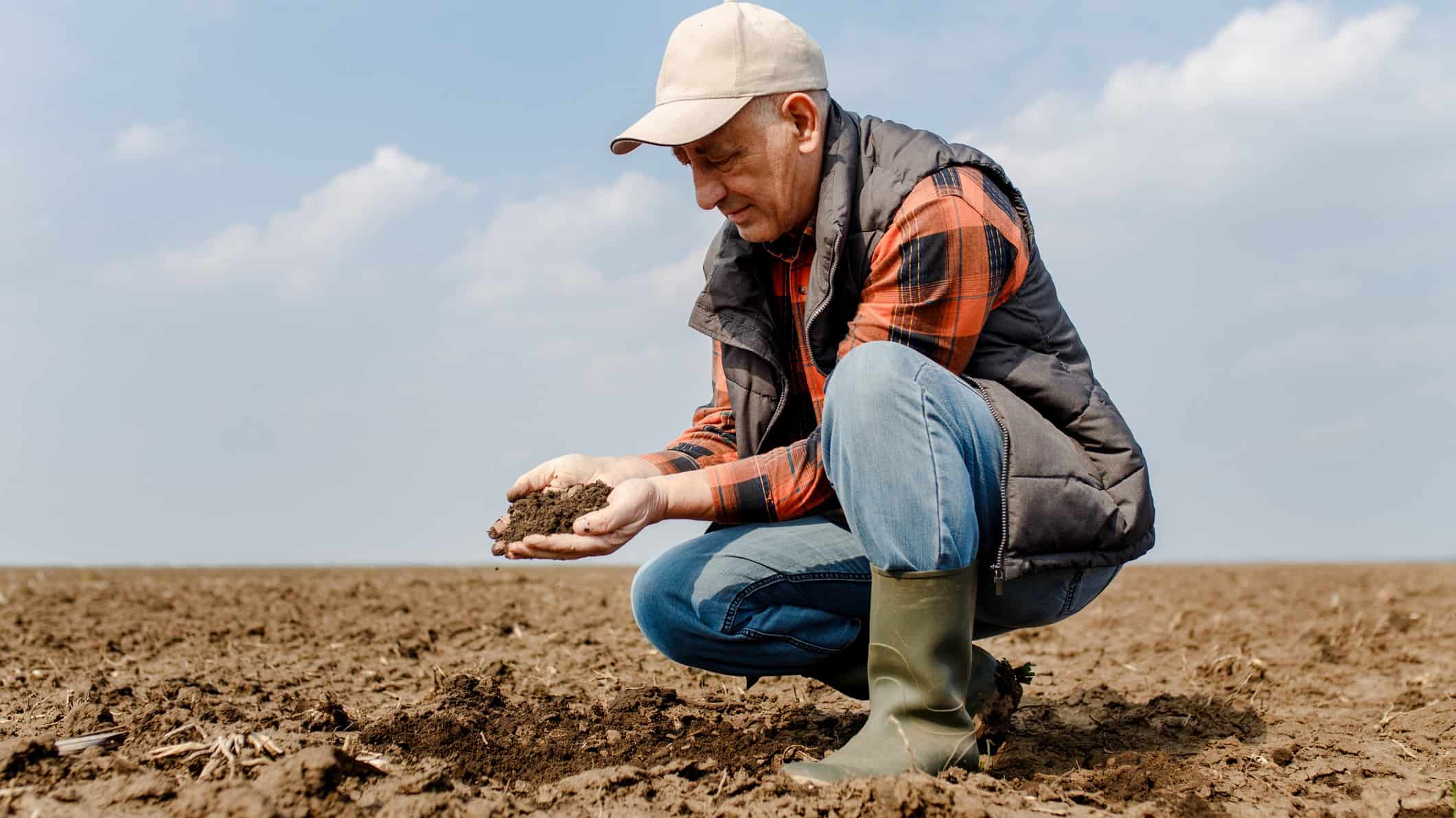 a farmer kneels on one leg and closely examines soil from his farm against a blue sky backdrop.