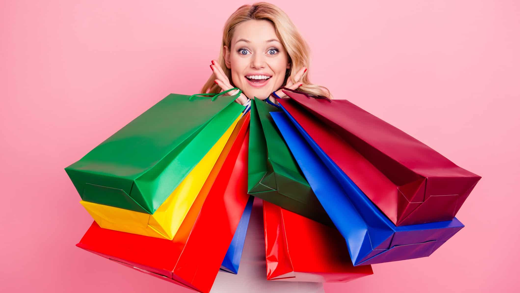 a woman smiles over the top of multiple shopping bags she is holding in both hands up near her face.