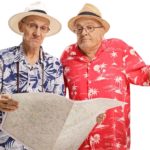 Two older men wearing colourful tropical patterned shirts and hats like tourists puzzle over a map one is holding.