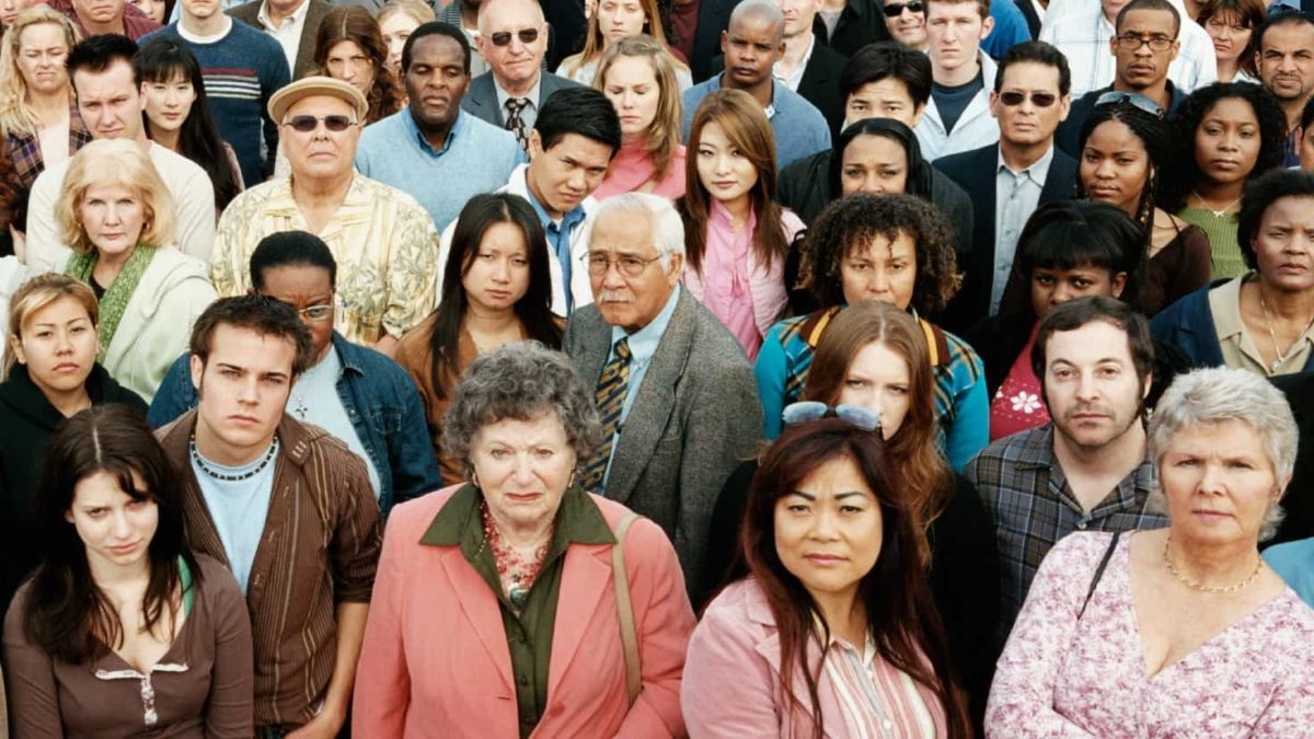 a diverse groups of about twenty people stand together in a crowd staring to the front with angry and annoyed looks on their faces.