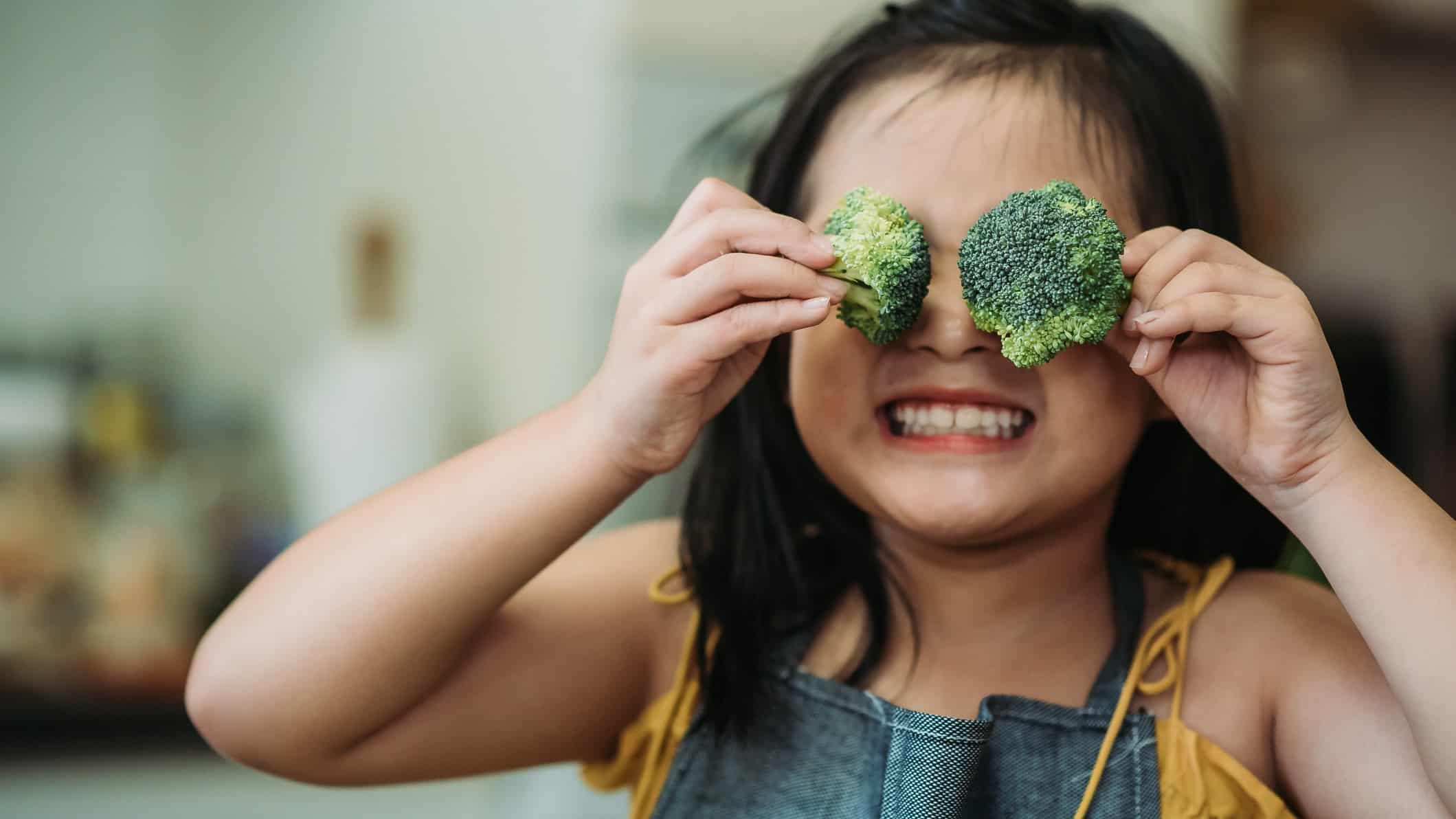 A little girl holds broccoli over her eyes with a big happy smile.
