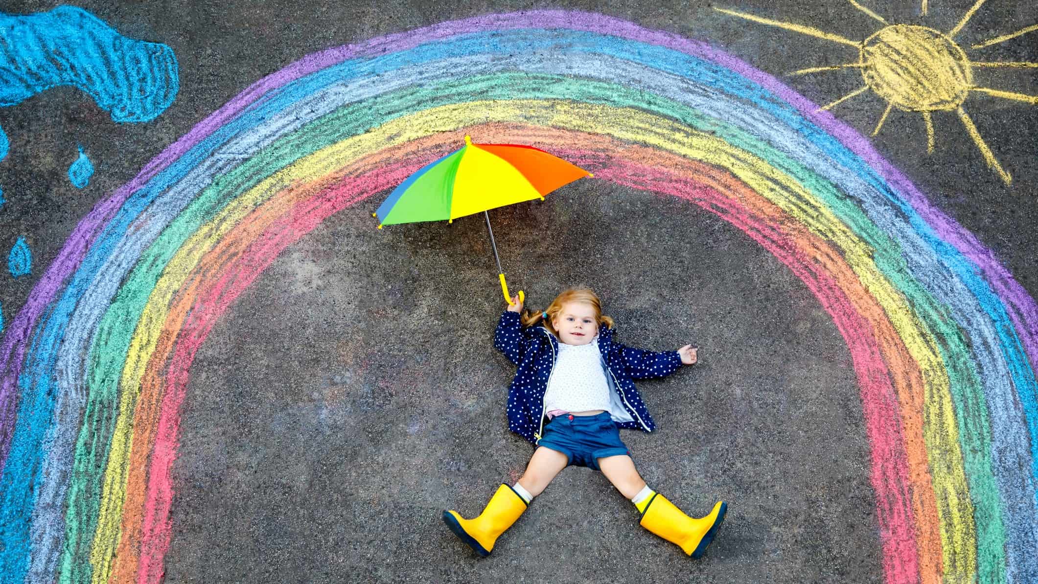 A little girl holds a colourful umbrella under a chalk rainbow drawn on the ground.