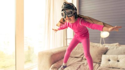A little girl dressed as a pilot prepares to leap off the sofa and take flight.