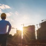 A man stands with hands on hips surveying construction of three high-rise buildings.