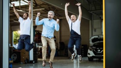 Three men leap for joy in front of a car dealership.