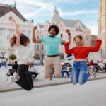 Three tourists jump high with big smiles in the village square.