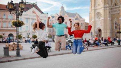 Three tourists jump high with big smiles in the village square.