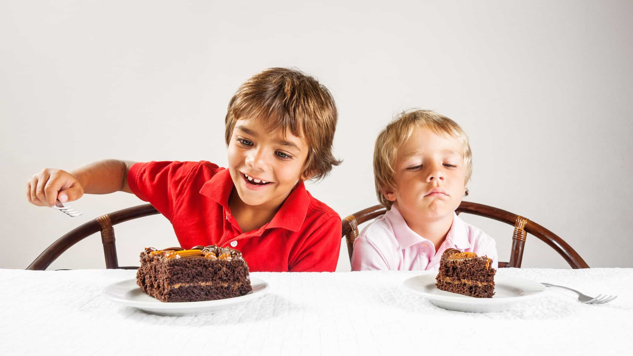 Two young boys each have a piece of chocolate cake, but one piece is bigger than the other.