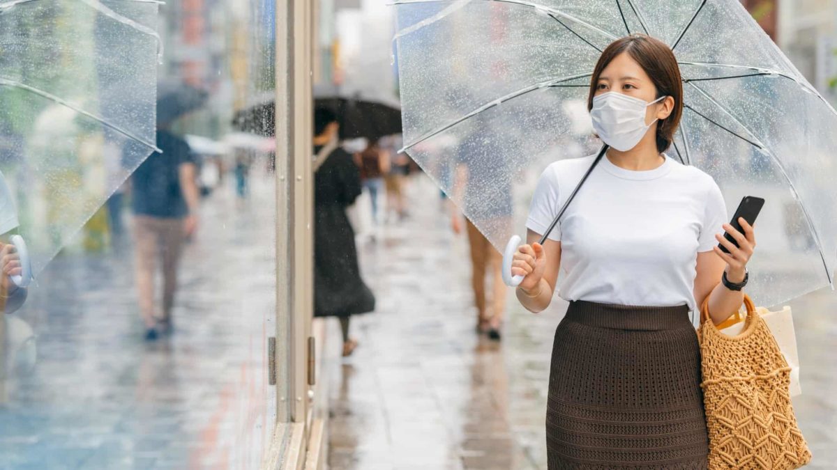A woman wearing a face mask and holding an umbrella, window shops in the rain.