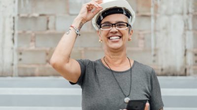 A woman wearing a hard hat and holding a device stands in front of a brick wall with a big smile on her face.