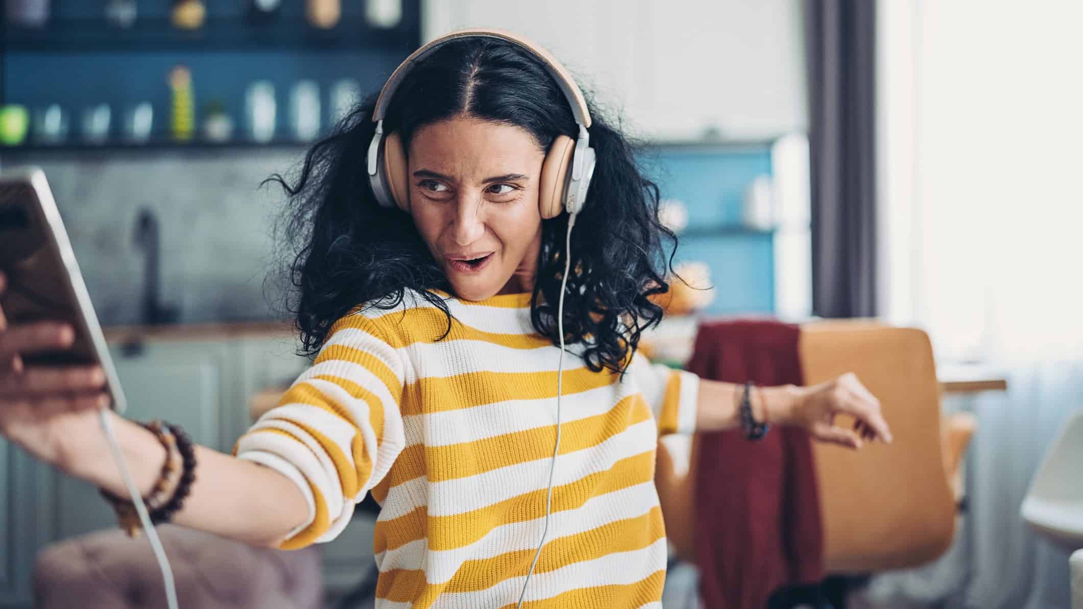 A woman wearing a yellow and white striped top and headphones plays excitedly with her phone.