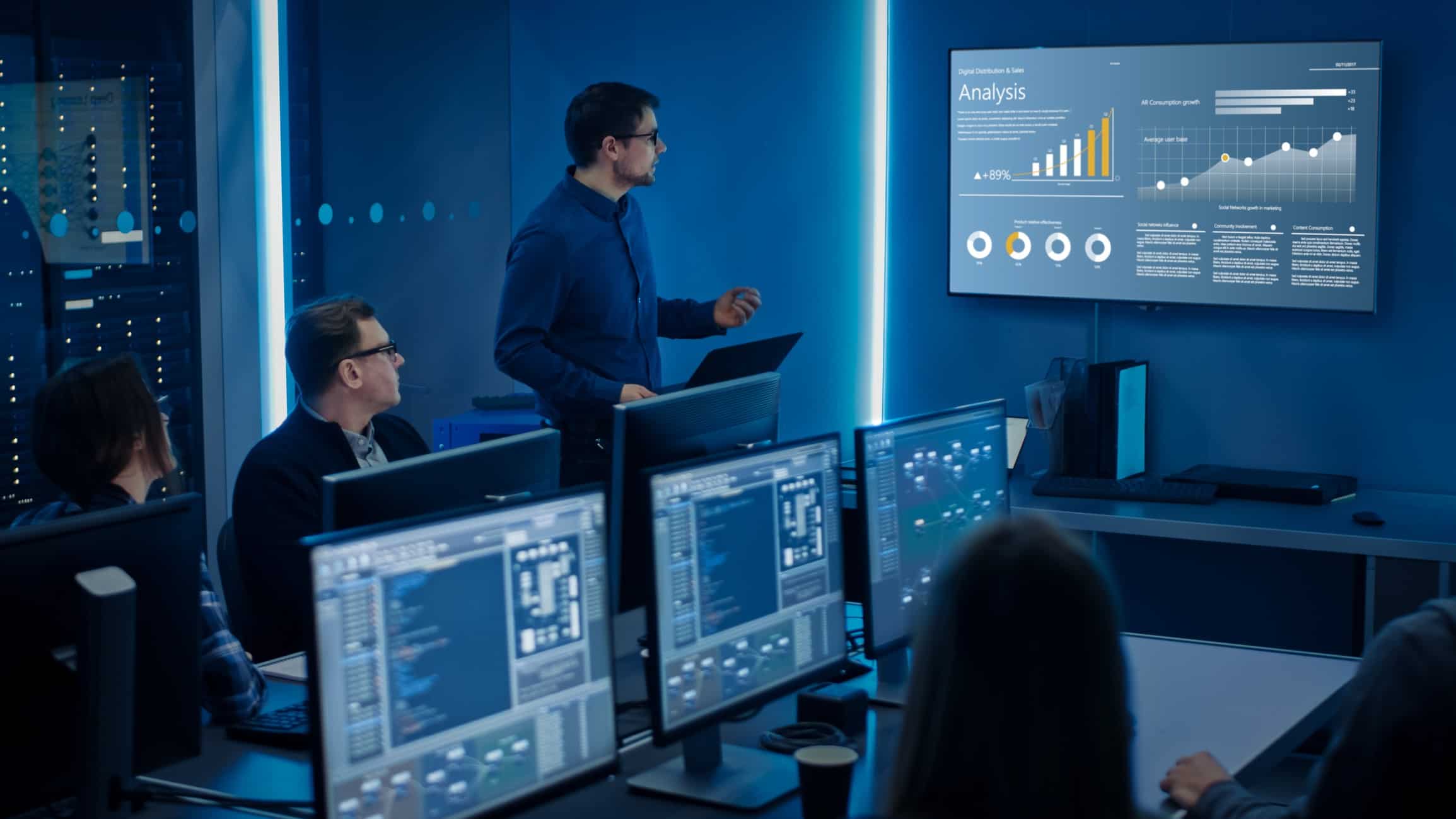 A group of executives sit in front of computer screens in a darkened room while a colleague stands giving a presentation with a share price graphic lit up on the wall