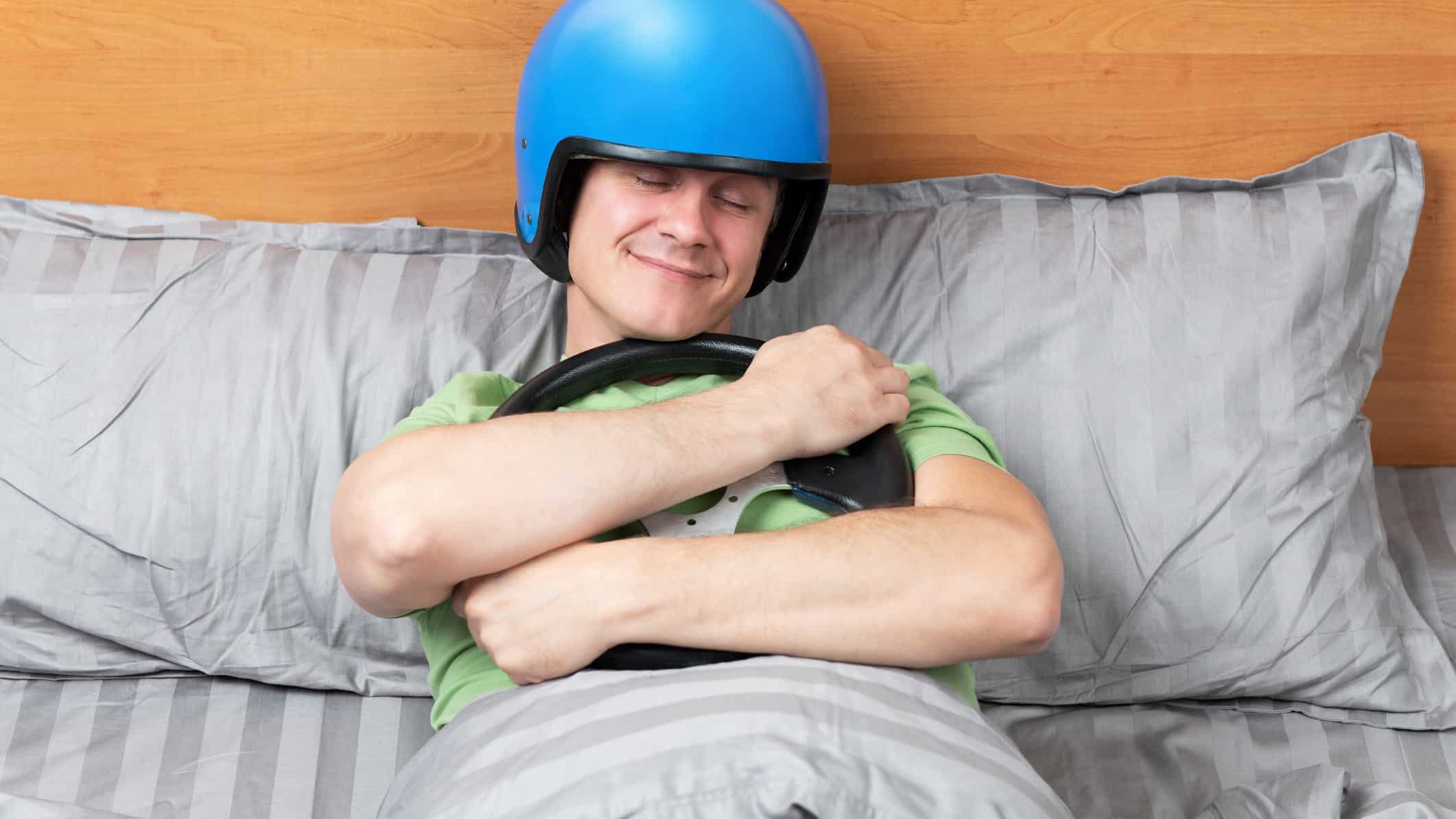A smiling man falls asleep wearing a racing helmet and hugging a steering wheel