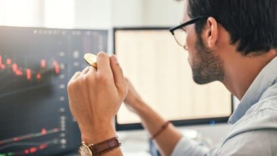 Man sitting at a desk facing his computer screen and holding a coin representing discussion by the RBA Governor about cryptocurrency and digital tokens