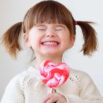 A little girl has a huge smile and a giant lollipop.