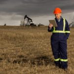 A Santos oil and gas company employee stands in a field looking at an ipad with an oil rig in the background and grey skies above representing carbon in the atmosphere
