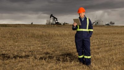 A Santos oil and gas company employee stands in a field looking at an ipad with an oil rig in the background and grey skies above representing carbon in the atmosphere