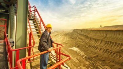 An engineer takes a break on a staircase and looks out over a huge open pit coal mine as the sun rises in the background.