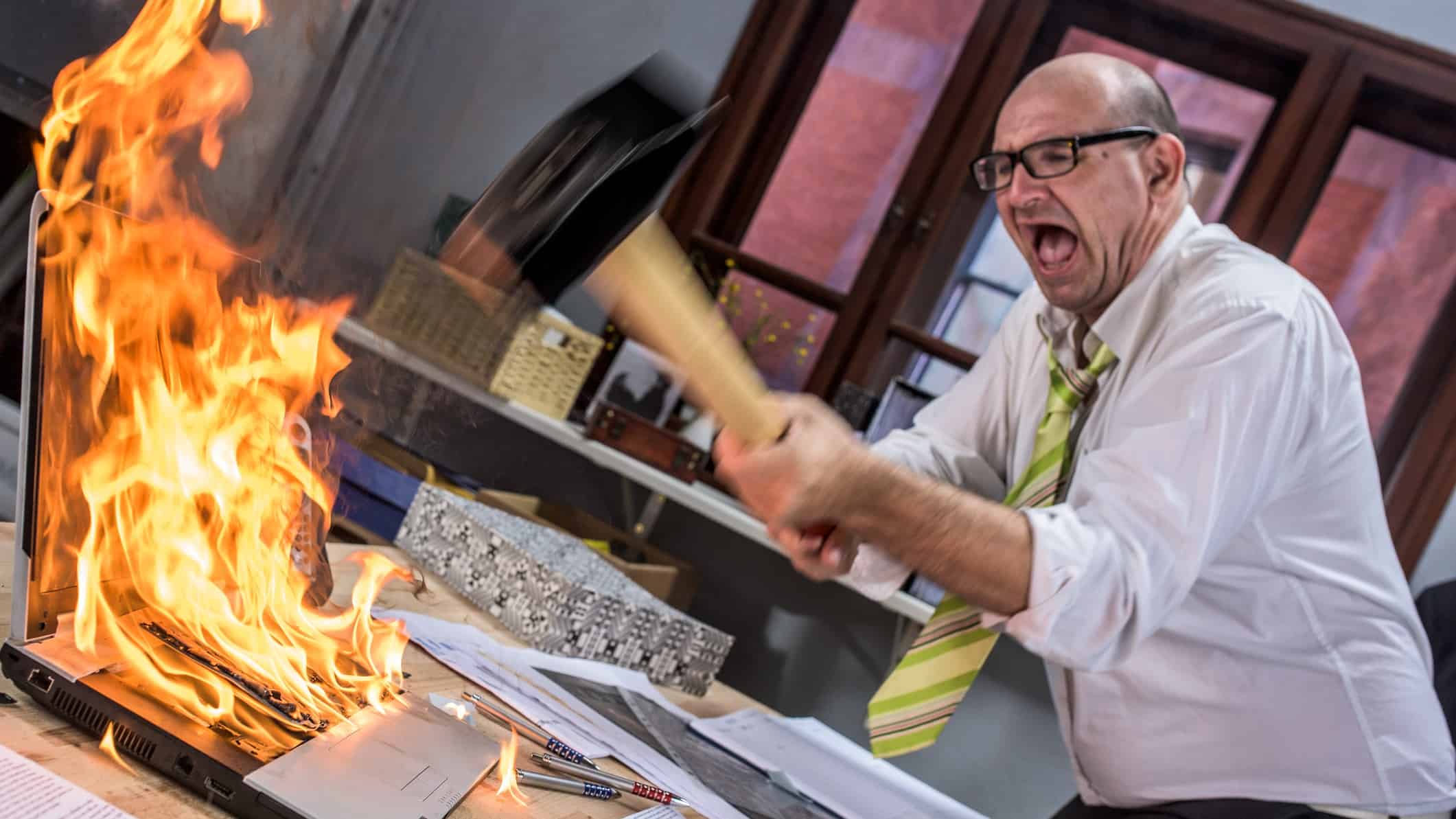 A businessman smashes his laptop with a hammer because it is on fire.
