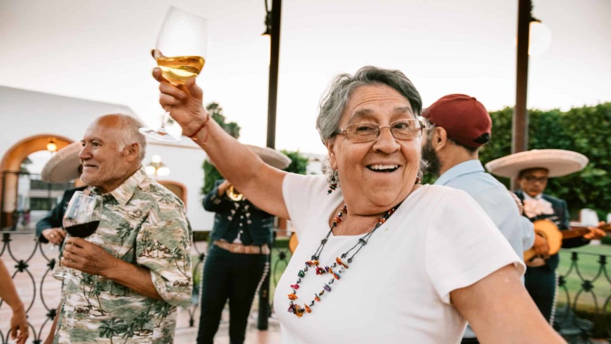 An elderly retiree holds her wine glass up while dancing at a party feeling happy about her ASX shares investments especially Brickworks for its dividends
