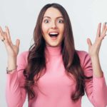 A wide-eyed happy woman with long brown hair and wearing a pink top holds her hands up in delight after hearing positive news
