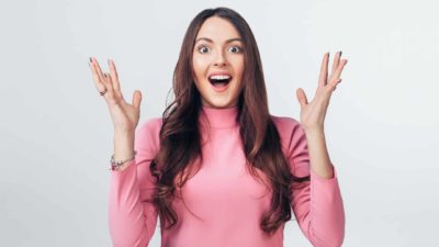 A wide-eyed happy woman with long brown hair and wearing a pink top holds her hands up in delight after hearing positive news