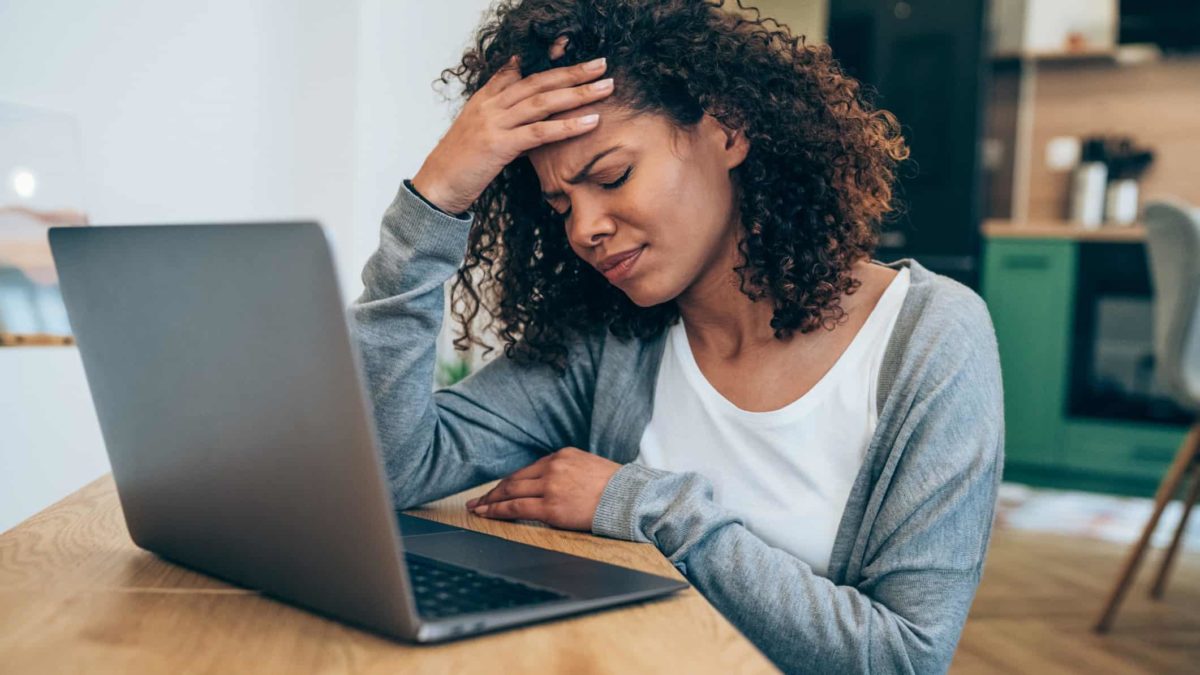 A disappointed female investor sits in front of her laptop and puts her hand to her forehead and closes her eyes in disappointment over share price falls