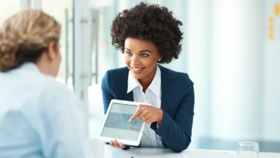 A female financial services professional with a manicured black afro hairstyle turns an ipad screen to show a client across the table a set of ASX shares figures in graph format.