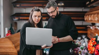 Two funeral workers with a laptop surrounded by cofins.