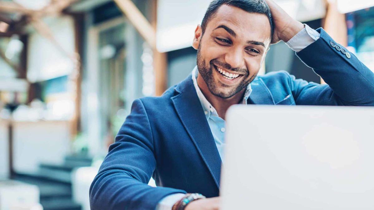 An executive in a suit smooths his hair and laughs as he looks at his laptop feeling surprised and delighted.