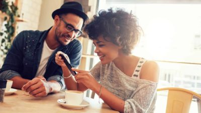 A happy man and woman sit having a coffee in a cafe while she holds up her phone to show him the ASX shares that did best today.