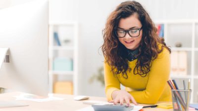 A young female investor with brown curly hair and wearing a yellow top and glasses sits at her desk using her calculator to work out how much her ASX dividend shares will pay this year